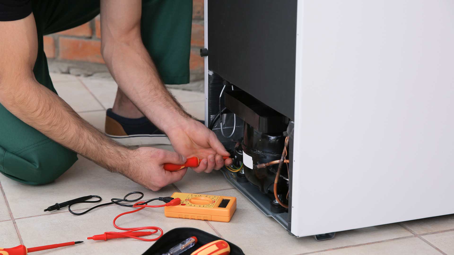 A man kneeling behind a refrigerator to tinker with the wiring behind it